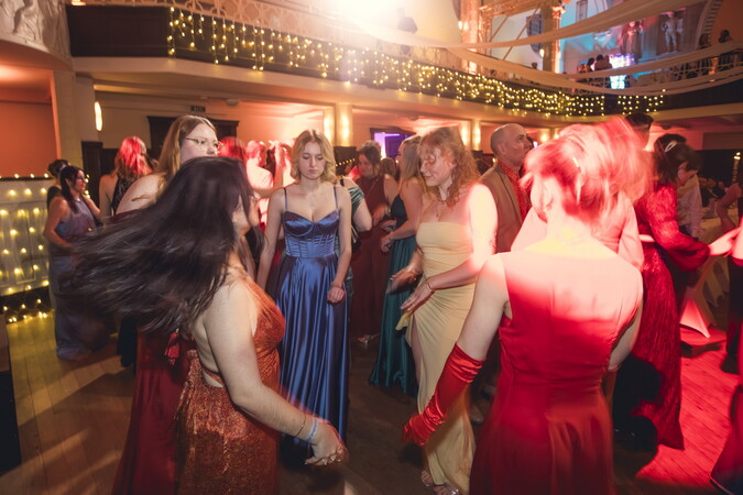 Tanzfläche in festlich geschmücktem Saal, auf der mehrere elegant gekleidete Personen in Abendgarderobe gemeinsam tanzen, umgeben von warmem Licht und Lichterketten auf der Galerie im Hintergrund.