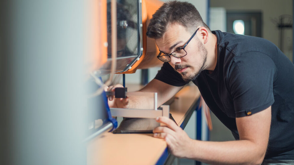 Eine Person mit Brille richtet konzentriert ein technisches Mess- oder Prüfgerät auf einer Werkbank aus. Sie beugt sich nach vorn und justiert eine Vorrichtung in einer hellen Werkstatt- oder Laborumgebung. Im Hintergrund sind weitere technische Geräte und Arbeitsflächen zu sehen.